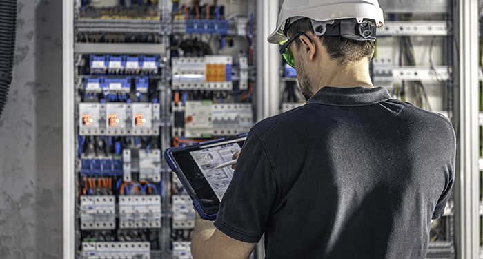 Man, an electrical working in a switchboard with fuses, uses a tablet.